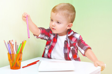 Cute little boy in a red checkered shirt smiles and draws a lilac marker in a white album.
