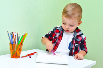 Cute little boy in a red checkered shirt smiles and draws a lilac marker in a white album
