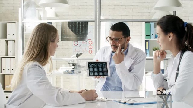 Zoom out shot of friendly black man in lab coat using tablet computer to show MRI images and explaining them to two female colleagues during meeting in clinic