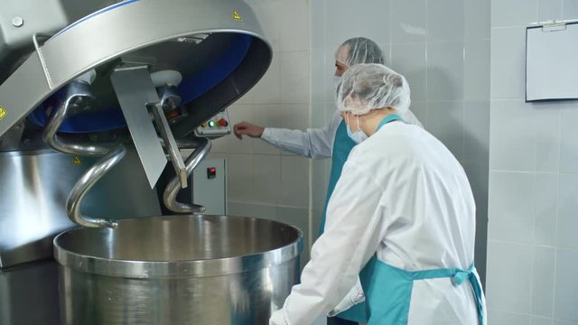 Two food production workers in protective wear, bouffant mob caps, and face masks operating industrial dough mixing machine when working at factory