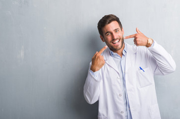 Handsome young professional man over grey grunge wall wearing white coat smiling confident showing and pointing with fingers teeth and mouth. Health concept.