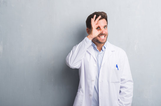Handsome young professional man over grey grunge wall wearing white coat doing ok gesture with hand smiling, eye looking through fingers with happy face.