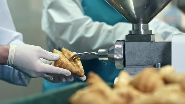 Tracking shot of two unrecognizable confectioners in gloves filling freshly baked croissants with cream