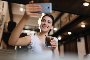 Beautiful woman using smartphone, taking selfie, drinking cocktail in cafe. Smiling blogger streaming video online 