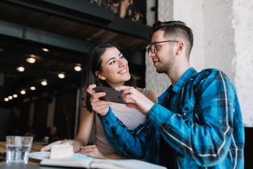 Stylish hipster couple making food photography, talking in loft cafe, have good mood. Cute woman with beautiful smile and her funny blogger friend communication together, holding mobile phone indoors