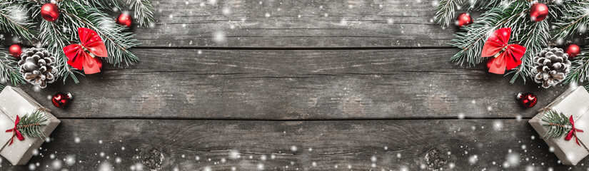 Panoramic Xmas theme with fir branches, red bows and globes, cones, present boxes, cookies and candies, on snowy rustic dark wooden background viewed from above, top, greeting card with space for text