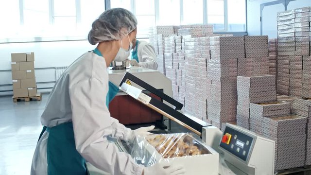 Woman in hairnet and face mask using factory machine when packing boxes with freshly baked cookies