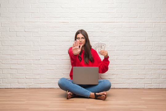 Young brunette woman sitting on the floor over white brick wall paying holding dollars with open hand doing stop sign with serious and confident expression, defense gesture