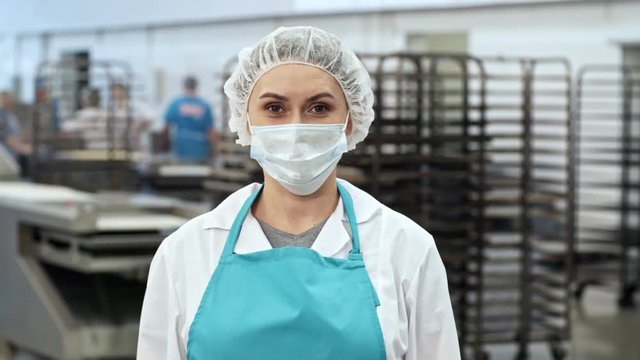 Tracking medium shot of cheerful female confectionery worker in apron, face mask and bouffant mob cap looking at camera