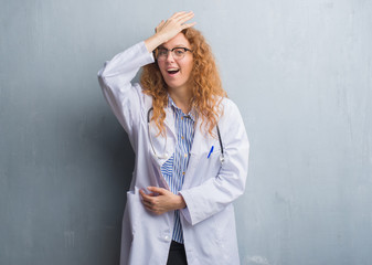 Young redhead doctor woman over grey grunge wall wearing a coat surprised with hand on head for...