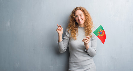 Young redhead woman over grey grunge wall holding flag of Portugal very happy pointing with hand and finger to the side