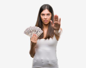 Young hispanic woman holding dollars with open hand doing stop sign with serious and confident expression, defense gesture