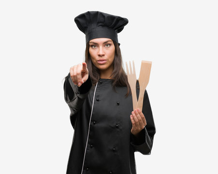 Young hispanic cook woman wearing chef uniform pointing with finger to the camera and to you, hand sign, positive and confident gesture from the front