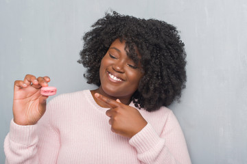 Young african american woman over grey grunge wall eating pink macaron very happy pointing with hand and finger
