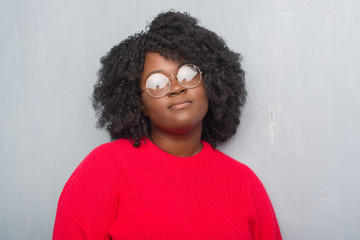 Young african american woman over grey grunge wall wearing winter sweater and glasses with serious...