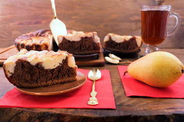 Pieces of chocolate cake with cottage cheese cut into pieces and laid out on a round plate, next to a ripe pear on a paper napkin and a glass of tea.