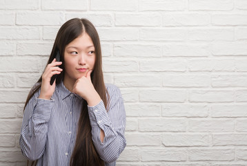 Young Chinese woman over brick wall talking on the phone serious face thinking about question, very confused idea