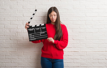 Young Chinese woman over white brick wall holding clapboard with a confident expression on smart face thinking serious