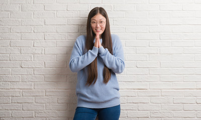 Young Chinise woman over white brick wall praying with hands together asking for forgiveness smiling confident.