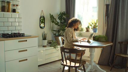 Charming young woman typing on laptop computer in a kitchen.