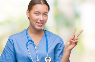 Young caucasian doctor woman wearing medical uniform over isolated background smiling with happy face winking at the camera doing victory sign. Number two.