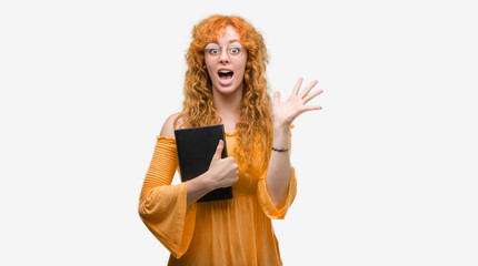 Young redhead student woman holding a book very happy and excited, winner expression celebrating victory screaming with big smile and raised hands