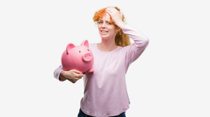 Young redhead woman holding piggy bank stressed with hand on head, shocked with shame and surprise face, angry and frustrated. Fear and upset for mistake.