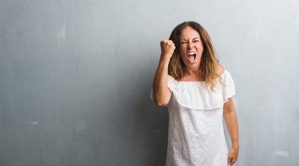 Middle age hispanic woman standing over grey grunge wall angry and mad raising fist frustrated and furious while shouting with anger. Rage and aggressive concept.