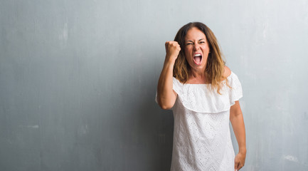 Fototapeta premium Middle age hispanic woman standing over grey grunge wall angry and mad raising fist frustrated and furious while shouting with anger. Rage and aggressive concept.