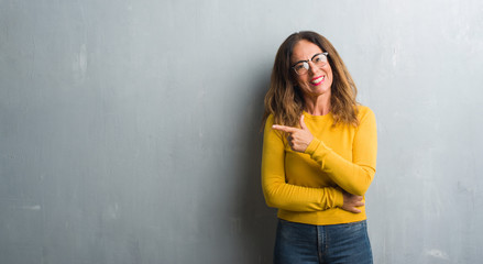 Fototapeta premium Middle age hispanic woman over grey wall wearing glasses cheerful with a smile of face pointing with hand and finger up to the side with happy and natural expression on face looking at the camera.