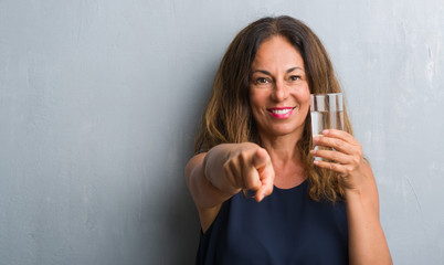 Middle age hispanic woman drinking glass of water pointing with finger to the camera and to you, hand sign, positive and confident gesture from the front