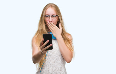 Blonde teenager woman holding smartphone and wearing headphones cover mouth with hand shocked with shame for mistake, expression of fear, scared in silence, secret concept