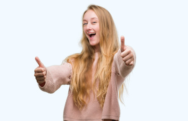 Blonde teenager woman wearing pink sweater approving doing positive gesture with hand, thumbs up smiling and happy for success. Looking at the camera, winner gesture.