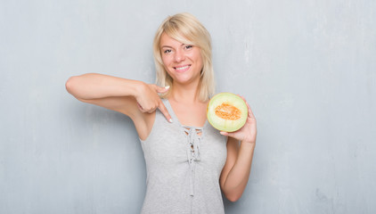 Adult caucasian woman over grunge grey wall eating cantaloupe melon with surprise face pointing finger to himself