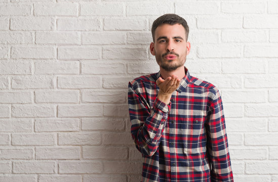 Young adult man standing over white brick wall looking at the camera blowing a kiss with hand on air being lovely and sexy. Love expression.