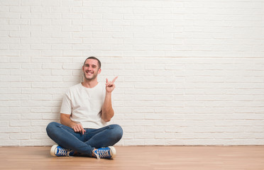 Young caucasian man sitting on the floor over white brick wall with a big smile on face, pointing with hand and finger to the side looking at the camera.