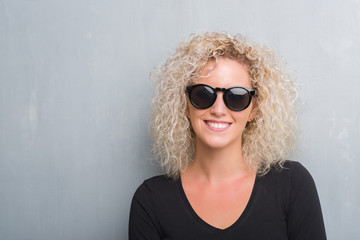 Young blonde woman with curly hair over grunge grey background with a happy and cool smile on face. Lucky person.