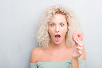 Young blonde woman over grunge grey background eating pink donut scared in shock with a surprise face, afraid and excited with fear expression