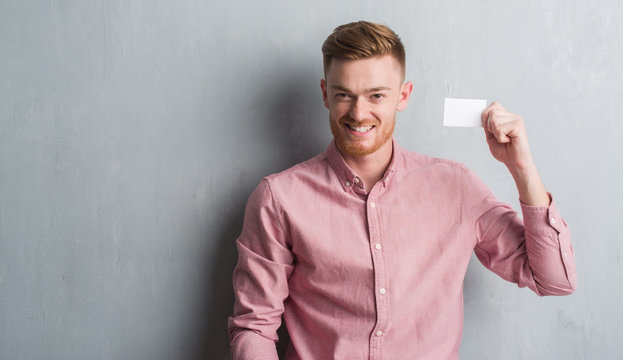 Young redhead man over grey grunge wall holding blank visit card with a happy face standing and smiling with a confident smile showing teeth