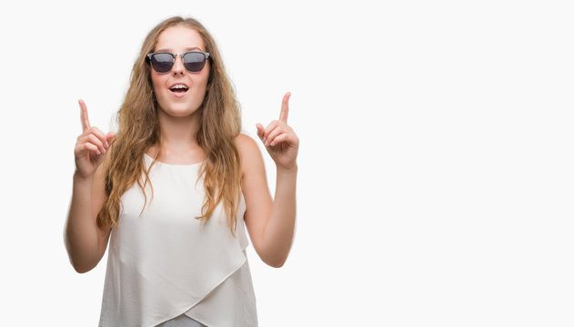 Young blonde woman wearing sunglasses amazed and surprised looking up and pointing with fingers and raised arms.