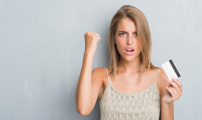 Fototapeta premium Beautiful young woman over grunge grey wall holding credit card annoyed and frustrated shouting with anger, crazy and yelling with raised hand, anger concept