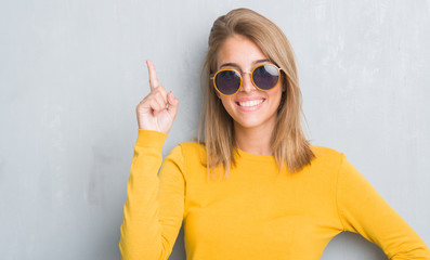 Beautiful young woman standing over grunge grey wall wearing retro sunglasses surprised with an idea or question pointing finger with happy face, number one