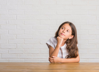 Young hispanic kid sitting on the table at home with hand on chin thinking about question, pensive expression. Smiling with thoughtful face. Doubt concept.