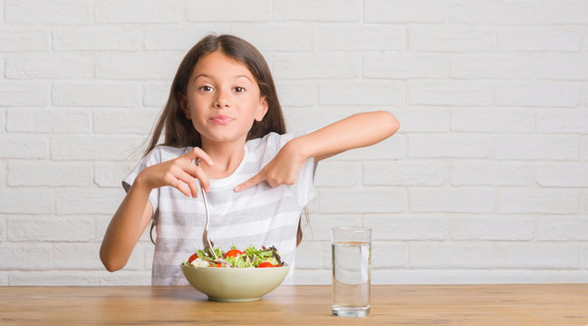 Young Hispanic Kid Sitting On The Table Eating Healthy Salad With Surprise Face Pointing Finger To Himself