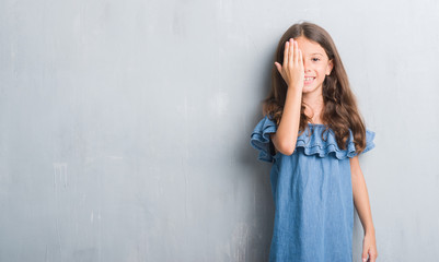 Fototapeta premium Young hispanic kid over grunge grey wall covering one eye with hand with confident smile on face and surprise emotion.