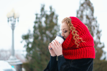 Girl with red hair in fur coat and red knitted scarf holds a mug of hot beverage in her hands and...