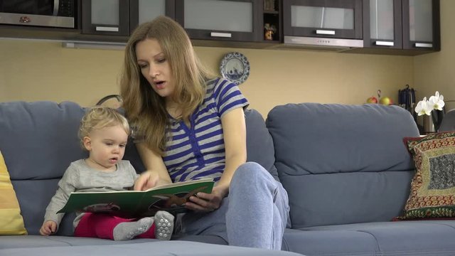 mother read book with little baby daughter on sofa in room. 4K