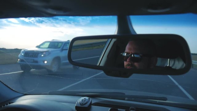 Young man in sunglasses is driving a car. Reflection face in rearview mirror of vehicle.