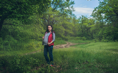 Naklejka na ściany i meble Portrait of a young beautiful pregnant brunette girl who walks through the green forest, wearing jeans and a red vest.