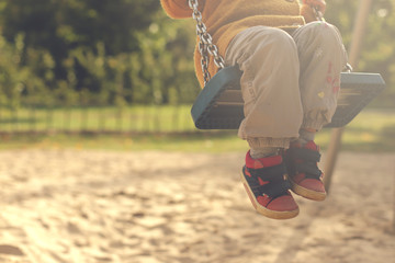 Child having fun with swing on a playground in bright afternoon sun - legs angled - matte look - obrazy, fototapety, plakaty